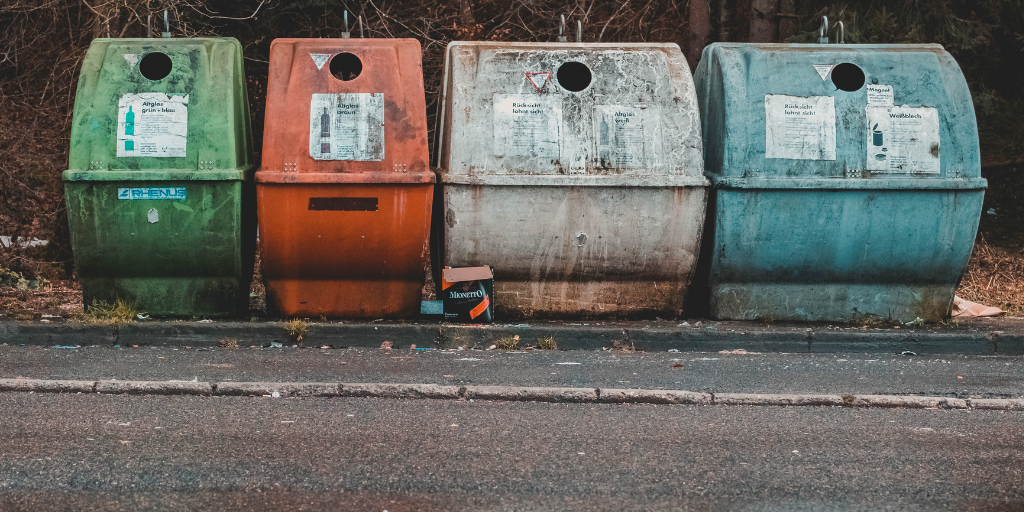 Four weathered recycling bins in green, orange, gray and blue along a roadside with a discarded cardboard box