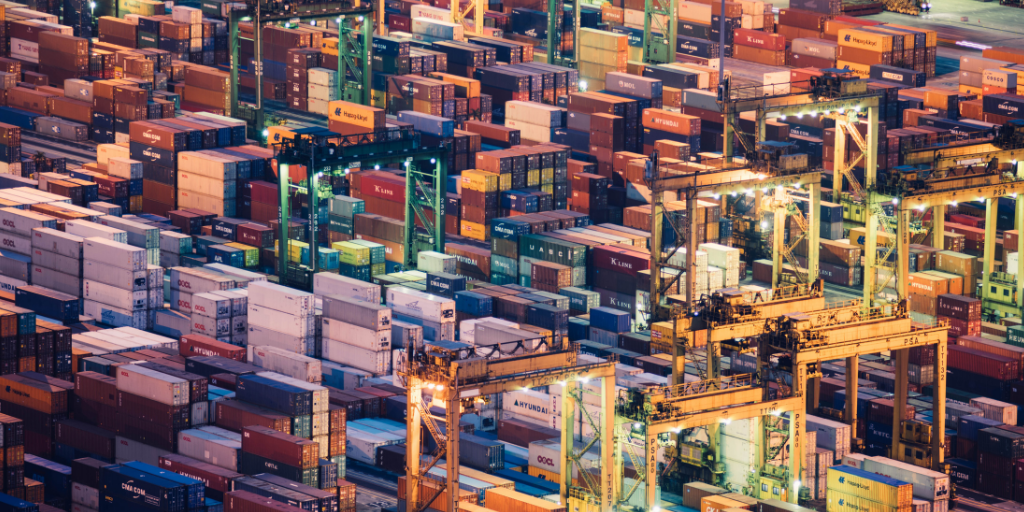 Shipping containers stacked at a busy port with gantry cranes loading containers at dusk