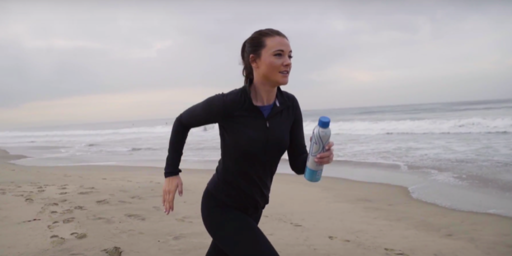 Woman running on a beach holding a PATH Water bottle with ocean waves in the background