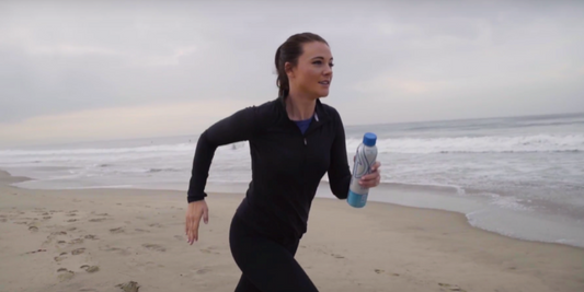 Woman running on a beach holding a PATH Water bottle with ocean waves in the background