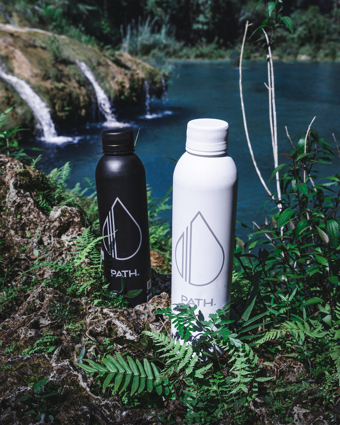 Front view of two PATH aluminum water bottles, black and white, showing droplet logo and caps by a blue river and waterfall