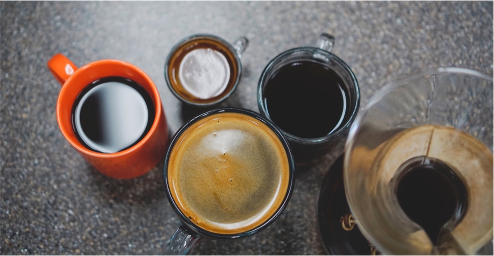 Overhead of coffee cups and carafe: black coffee in an orange mug, espresso with crema, and brewed coffee on a speckled counter