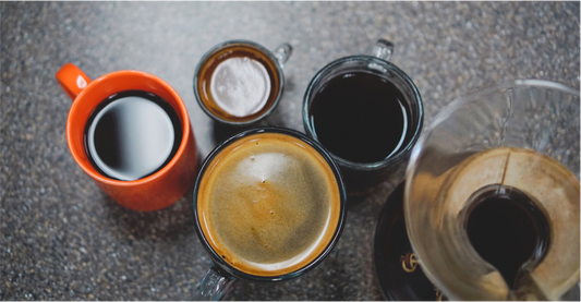 Overhead of coffee cups and carafe: black coffee in an orange mug, espresso with crema, and brewed coffee on a speckled counter