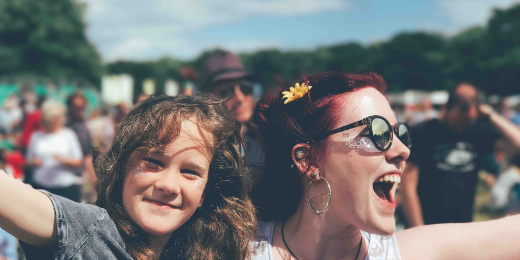 Young woman cheering and girl smiling at an outdoor music festival, both wearing glitter with a crowd and trees in the background