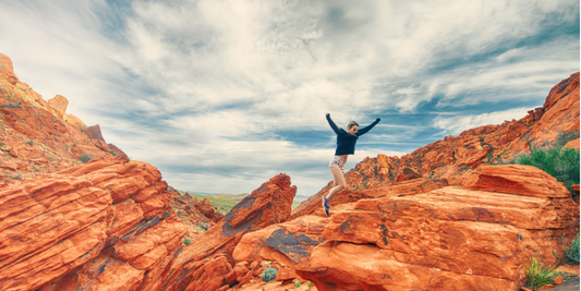 Person jumping between red sandstone rock formations in a desert landscape under a dramatic cloudy sky