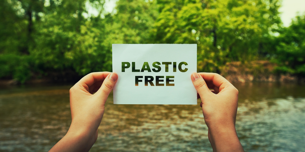 Two hands holding a card that reads PLASTIC FREE in front of a river and green trees