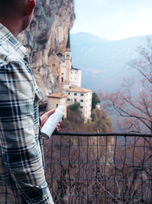 Person holding white PATH aluminum water bottle at a cliffside overlook with church and misty mountains in the background