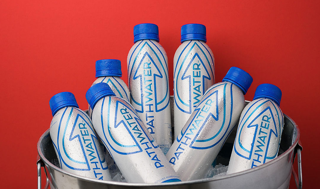 Front-angled view of PATHWATER aluminum water bottles with blue caps in an ice-filled metal bucket against a red background