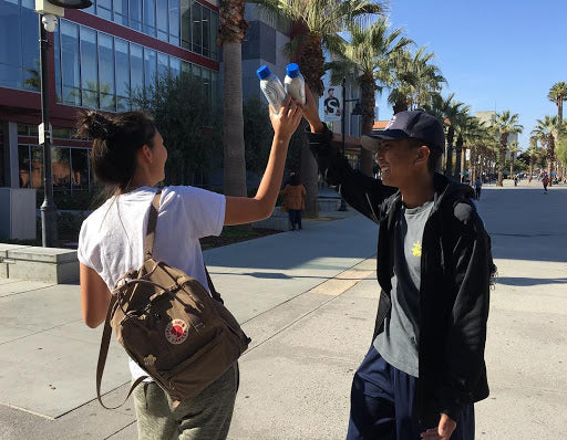Two students clinking reusable water bottles on a sunny college campus walkway
