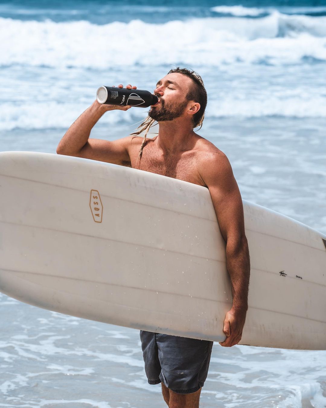 Surfer drinking from a black PATH aluminum bottle while holding a white surfboard with ocean waves in the background