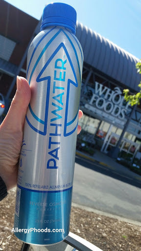 Close-up of PATHWATER aluminum bottle with blue cap held in a hand, front label visible, Whole Foods storefront in background