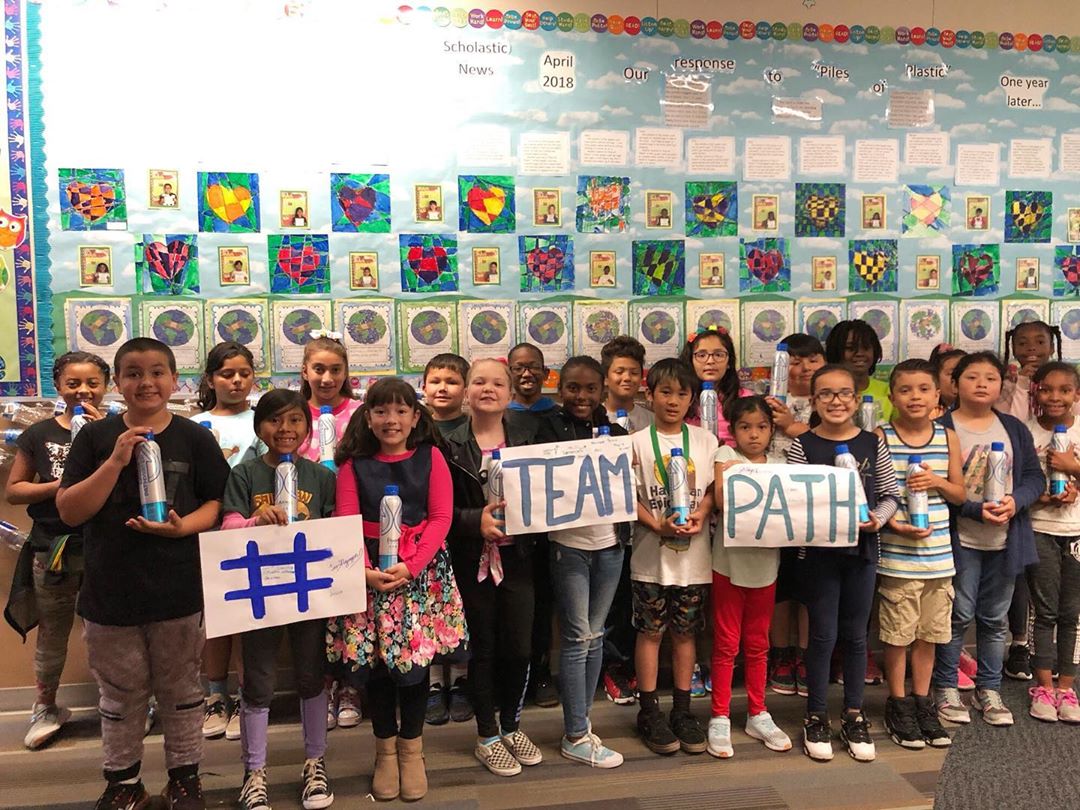 Students holding PATHWATER bottles and signs reading 'TEAM PATH' and a hashtag in front of classroom artwork