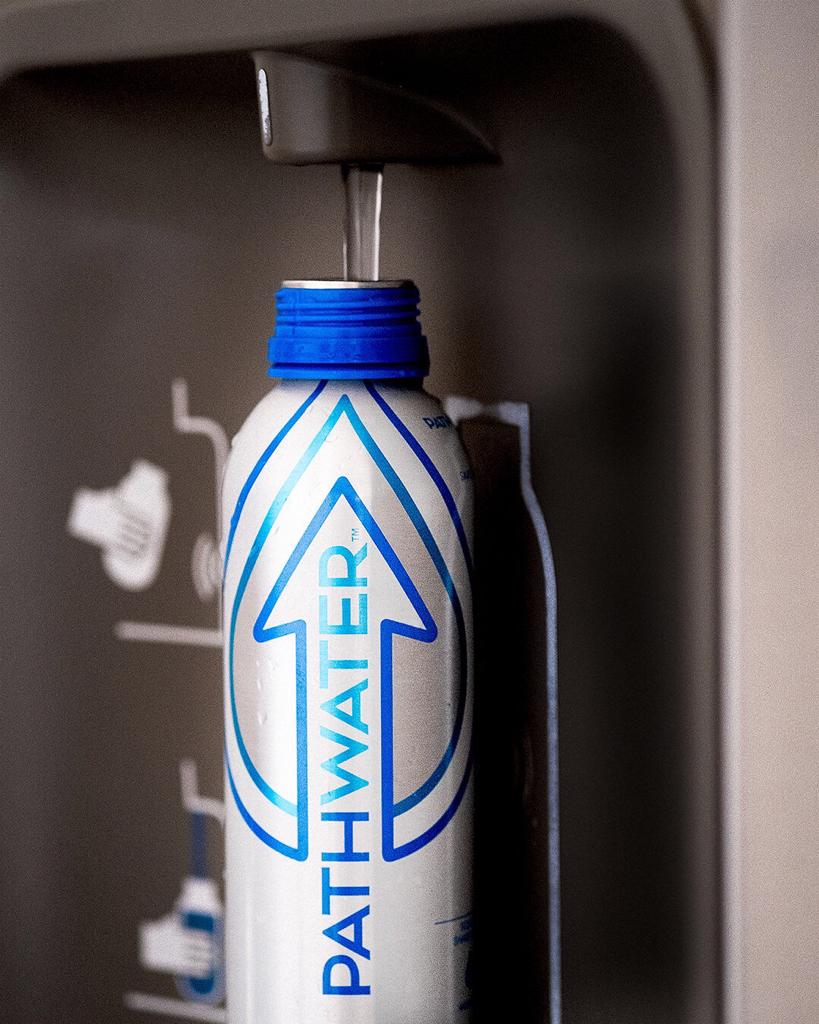Close-up front view of PATHWATER aluminum bottle showing blue arrow logo and filling water from a dispenser