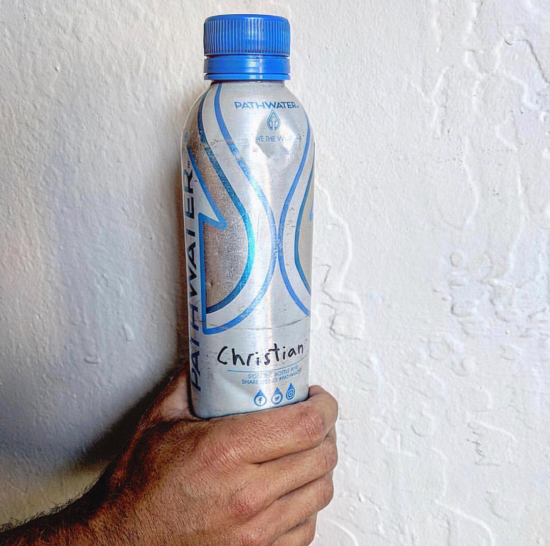 Front view of PATH Water aluminum bottle with blue cap showing handwritten name "Christian" held against a textured white wall