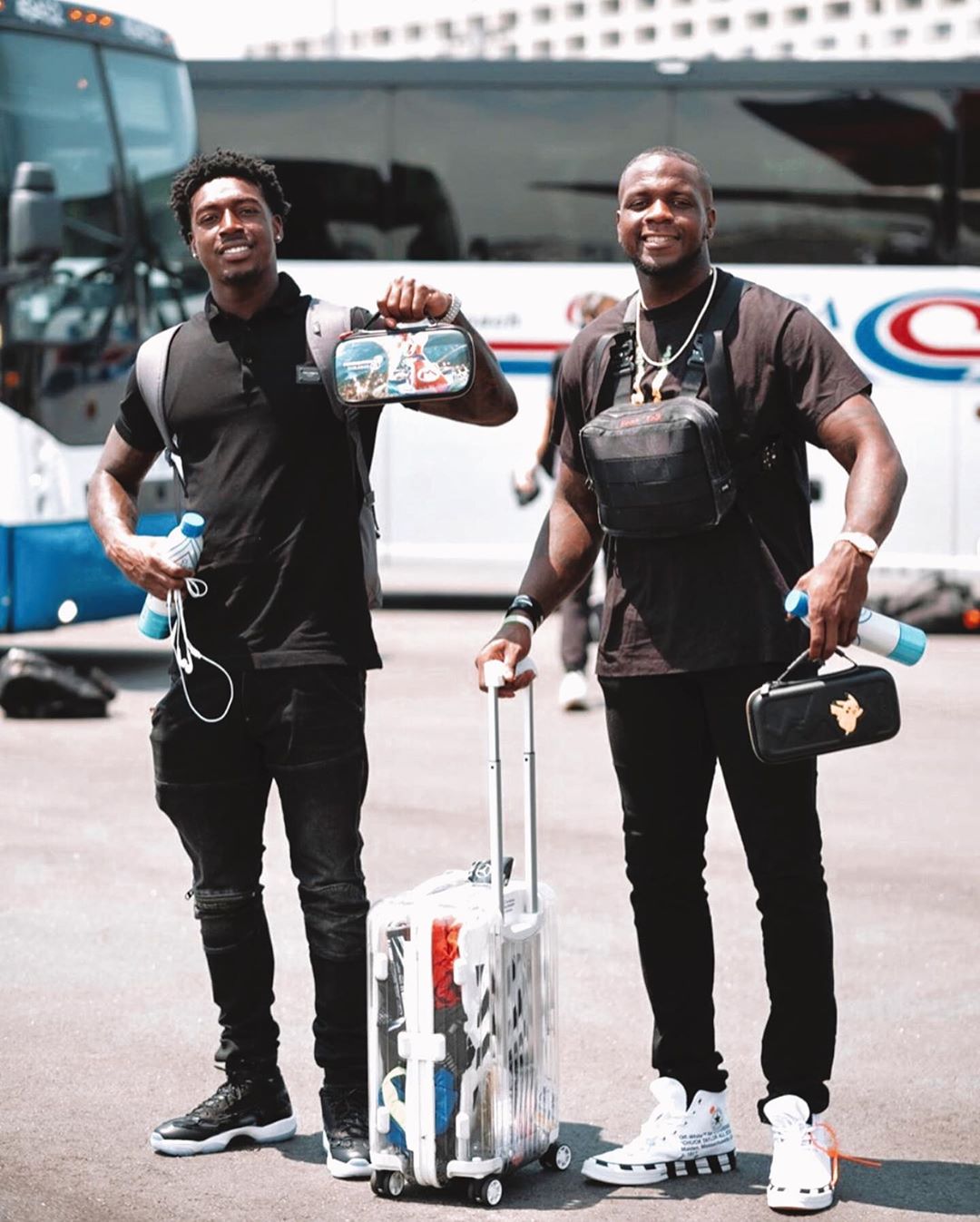 Two men standing on an airport tarmac holding reusable water bottles and travel bags next to a clear rolling suitcase