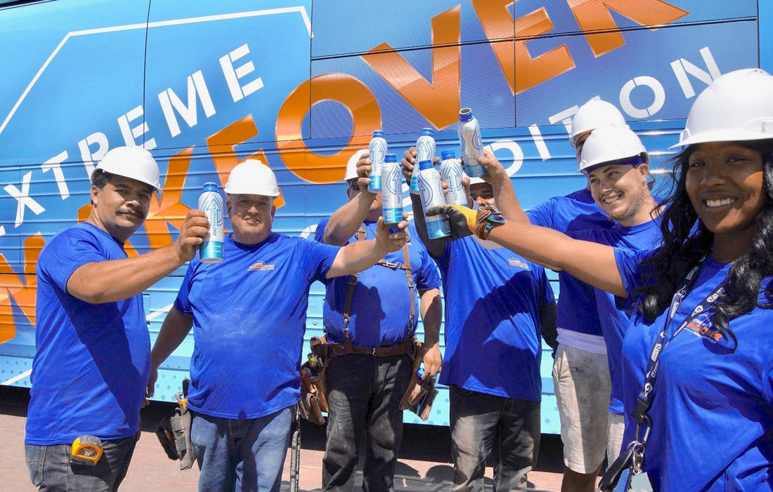 Construction workers in white hard hats and blue shirts raising PATHWATER aluminum bottles in front of a blue bus with 'EXTREME MAKEOVER' lettering