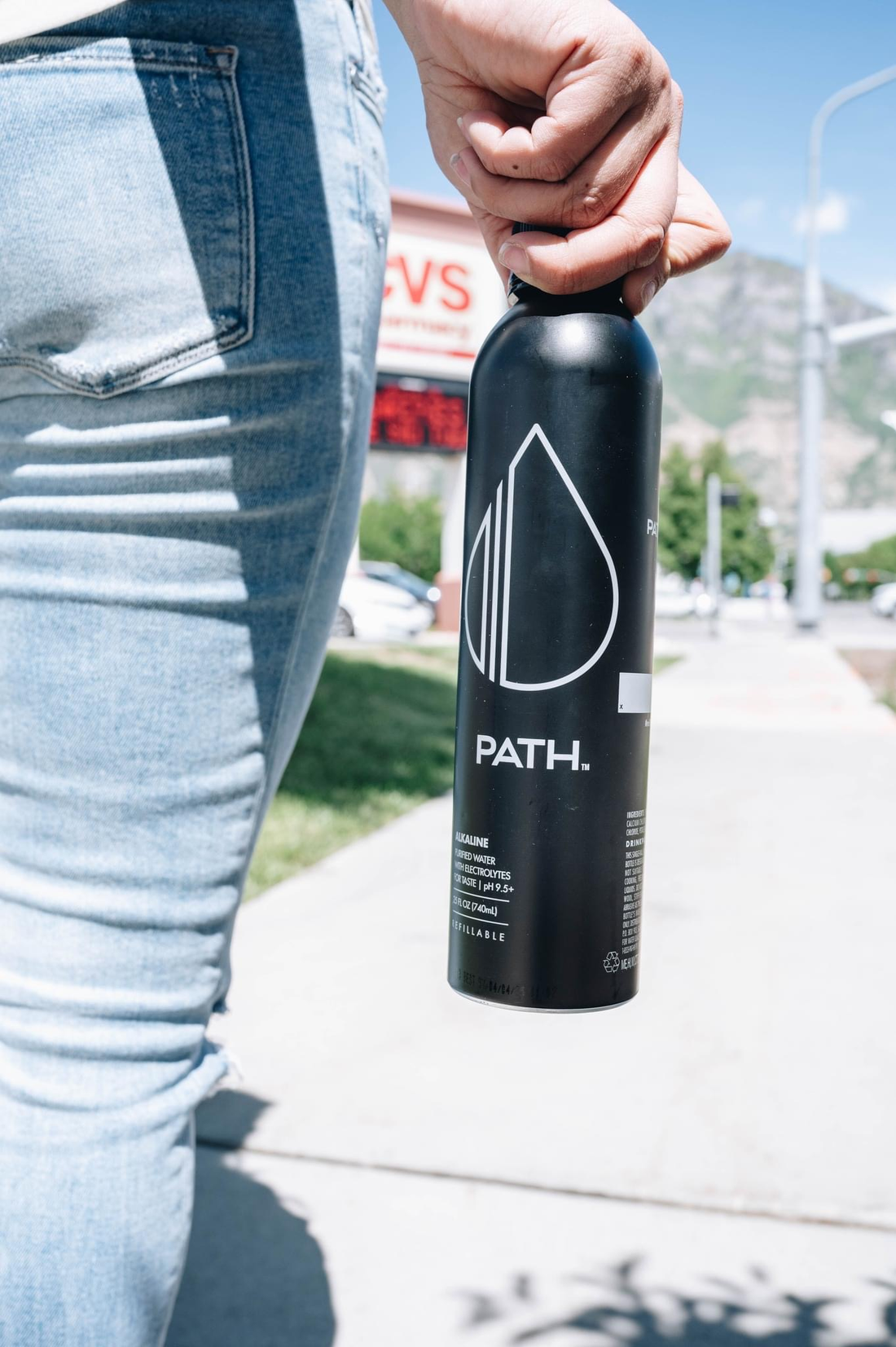 Side view of black PATH reusable water bottle held by hand showing PATH logo and drop design on a sunny sidewalk