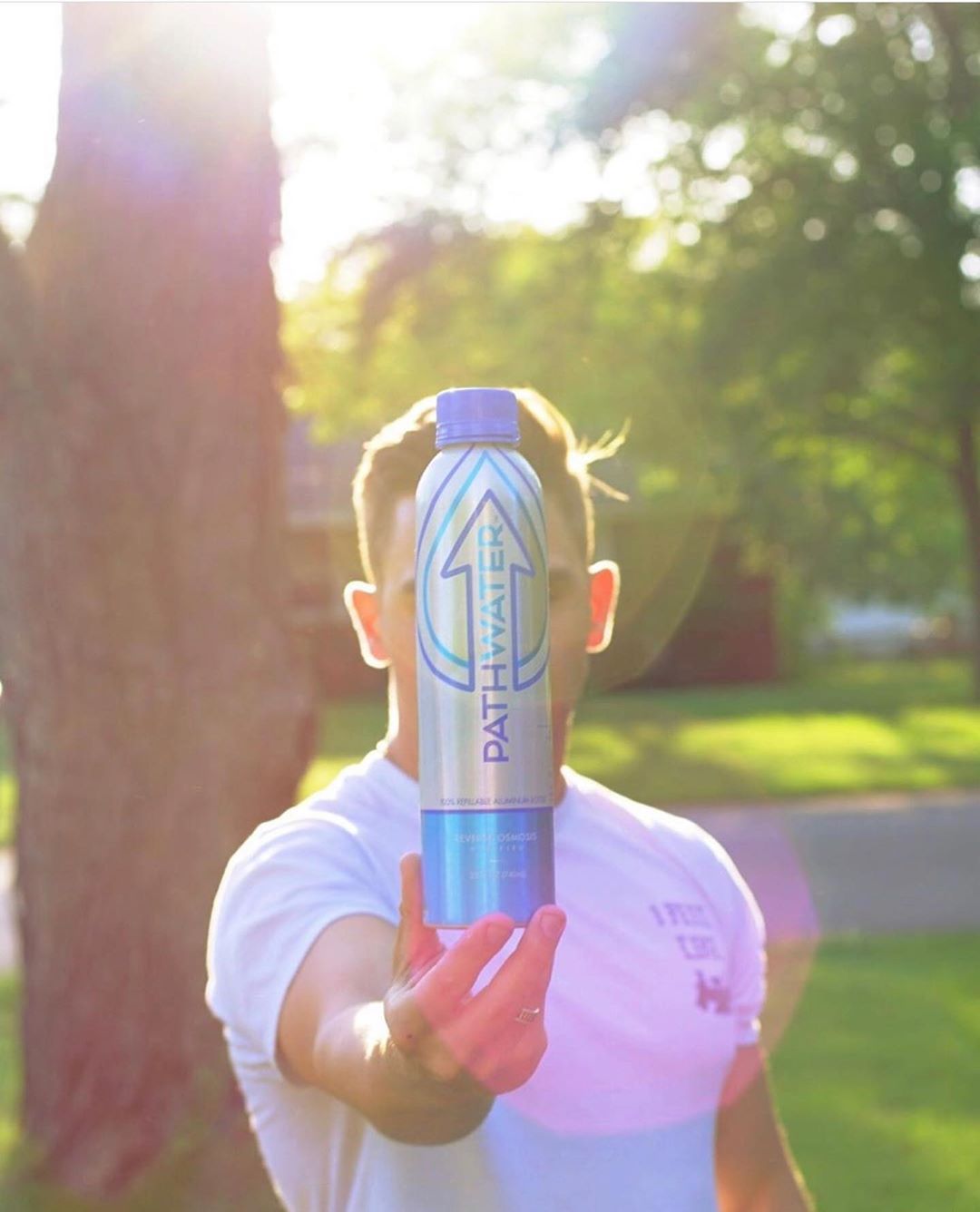 Front view of PATHWATER aluminum reusable bottle held at arm's length showcasing vertical logo and blue cap with sunlit park background
