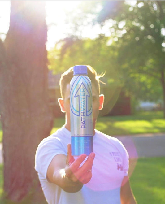 Front view of PATHWATER aluminum reusable bottle held at arm's length showcasing vertical logo and blue cap with sunlit park background
