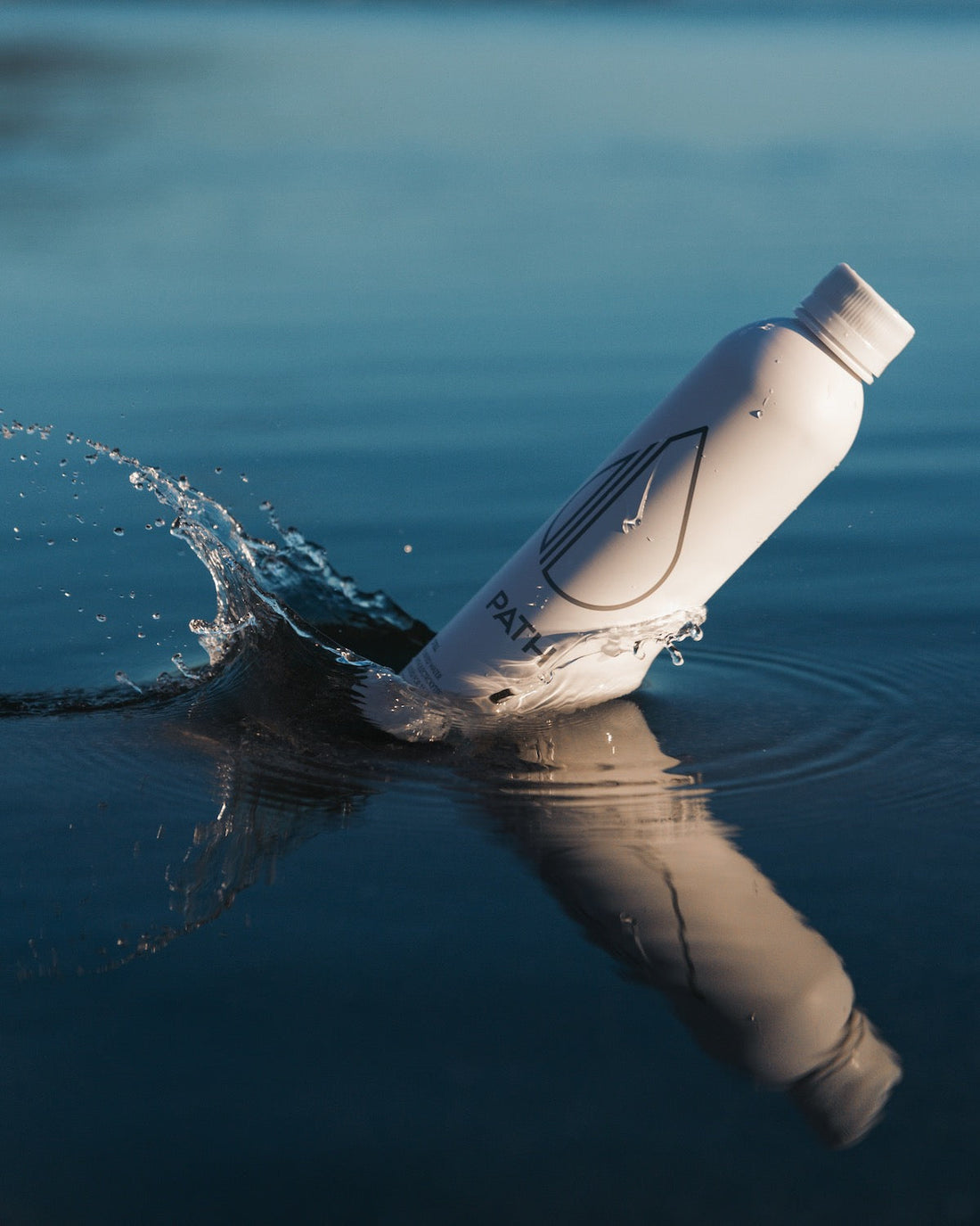 Angled view of white PATH aluminum water bottle showcasing the droplet logo and splashing on calm blue water