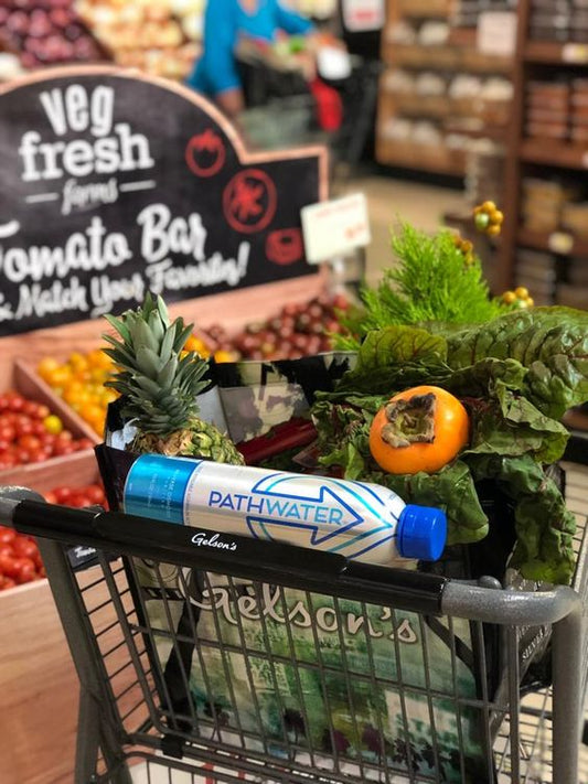 PATHWATER aluminum bottle resting in a shopping cart with pineapple, leafy greens, persimmon, and tomatoes in a grocery store produce aisle
