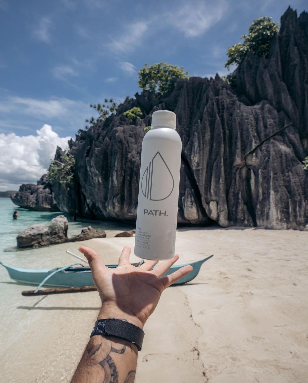 Hand tossing a PATH aluminum water bottle on a sunny beach with limestone cliffs and a small blue boat