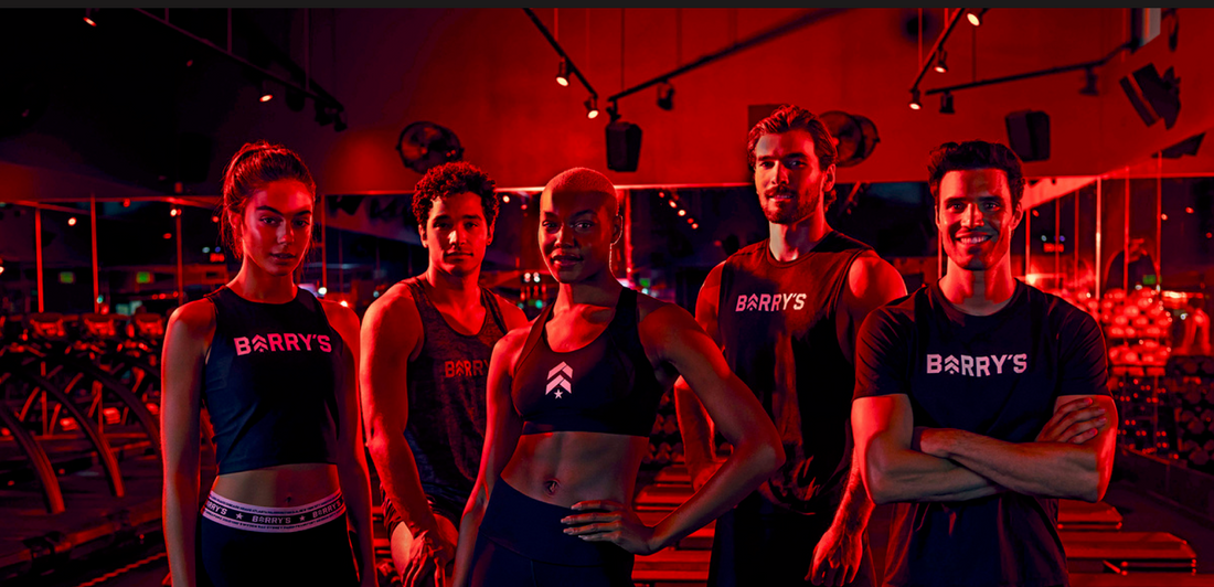 Five Barry's fitness instructors standing together in a red-lit Barry's studio with treadmills and mirrored walls