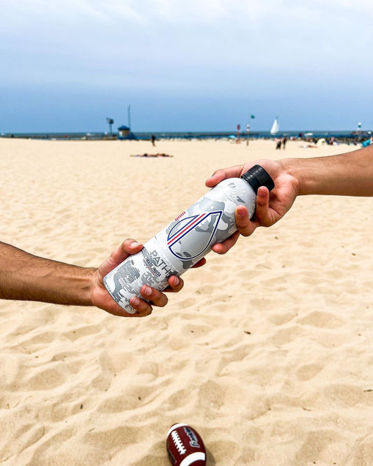 Two people passing a PATH aluminum water bottle on a sandy beach with the ocean and sailboats in the background