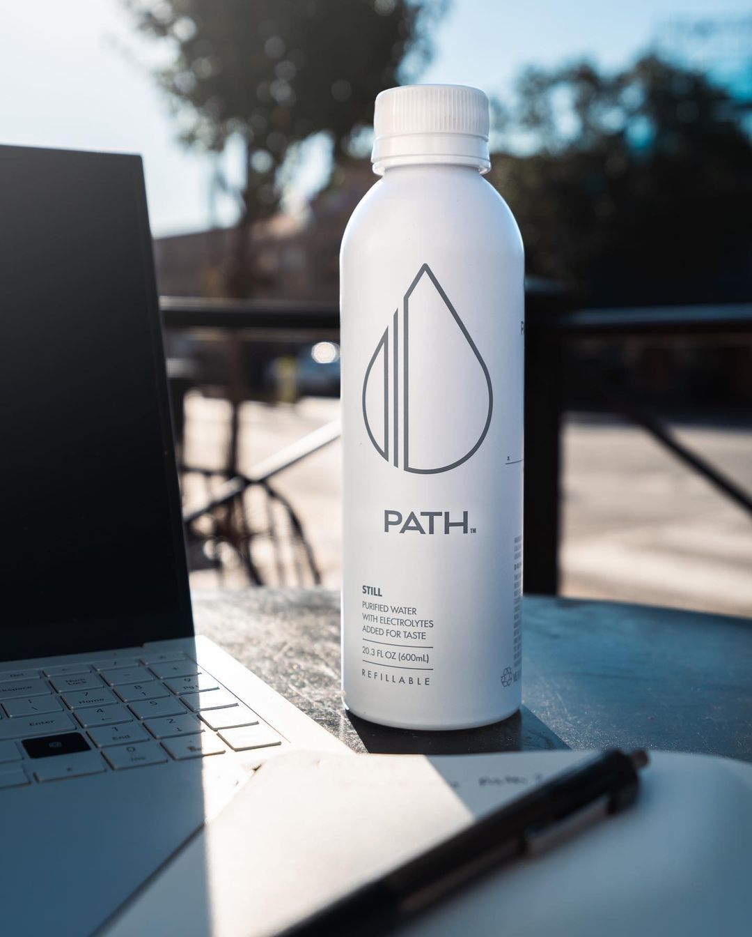 Front view of PATH white aluminum water bottle showing droplet logo with laptop and notebook on outdoor table