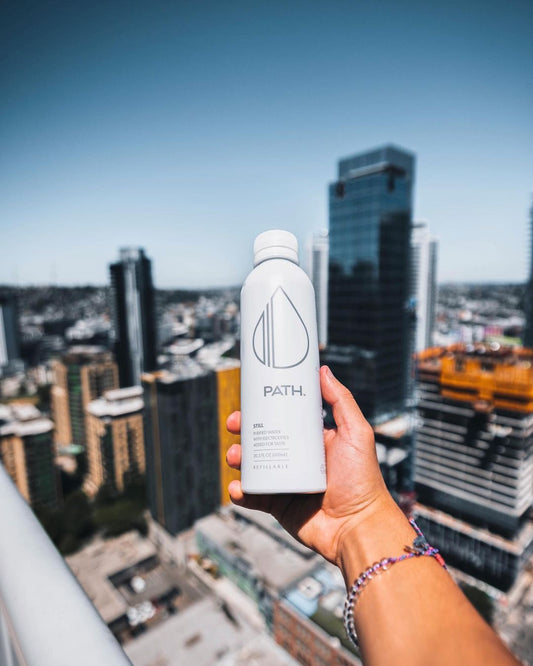 Hand holding a white PATH water bottle above a blurred urban skyline from a rooftop