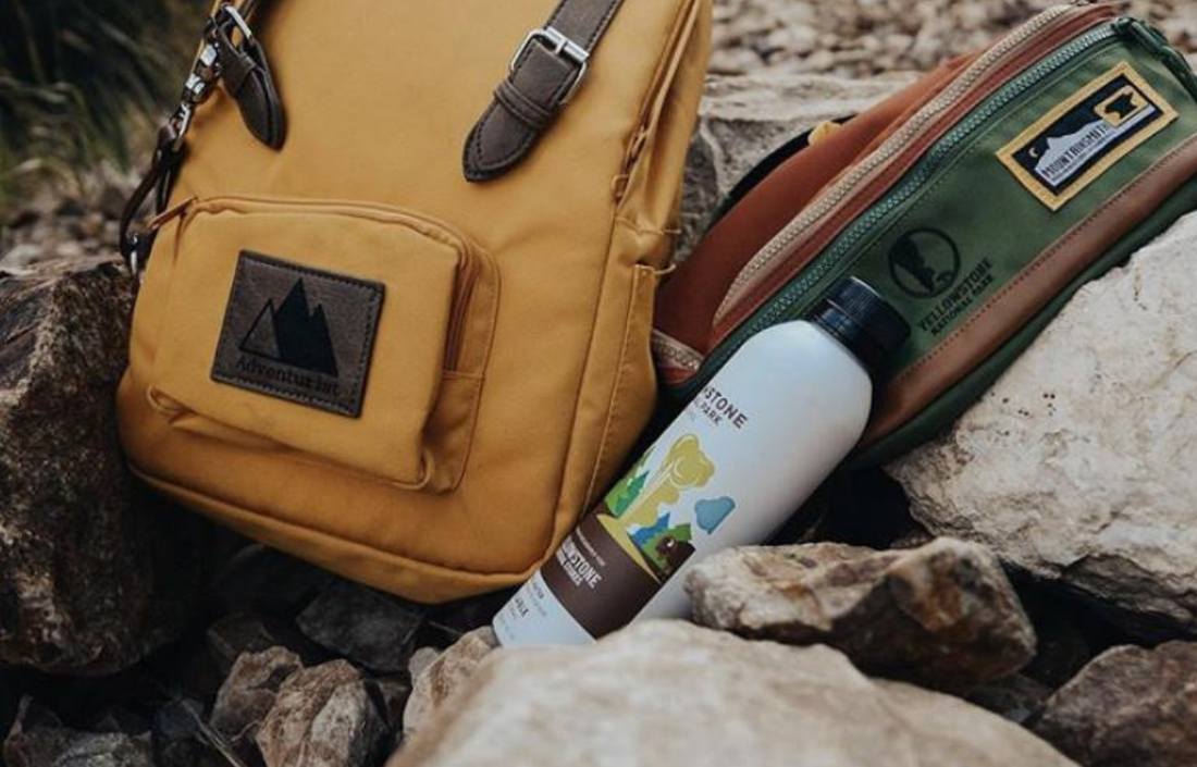 Angled view of PATH aluminum water bottle with Yellowstone co-branded label and black screw cap beside a yellow backpack on rocky ground