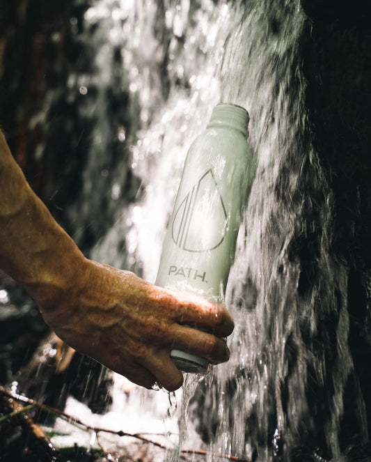 Hand holding a PATH aluminum water bottle under a waterfall