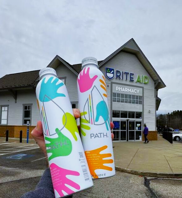 Two aluminum PATH water bottles held by a hand in front of a Rite Aid pharmacy storefront