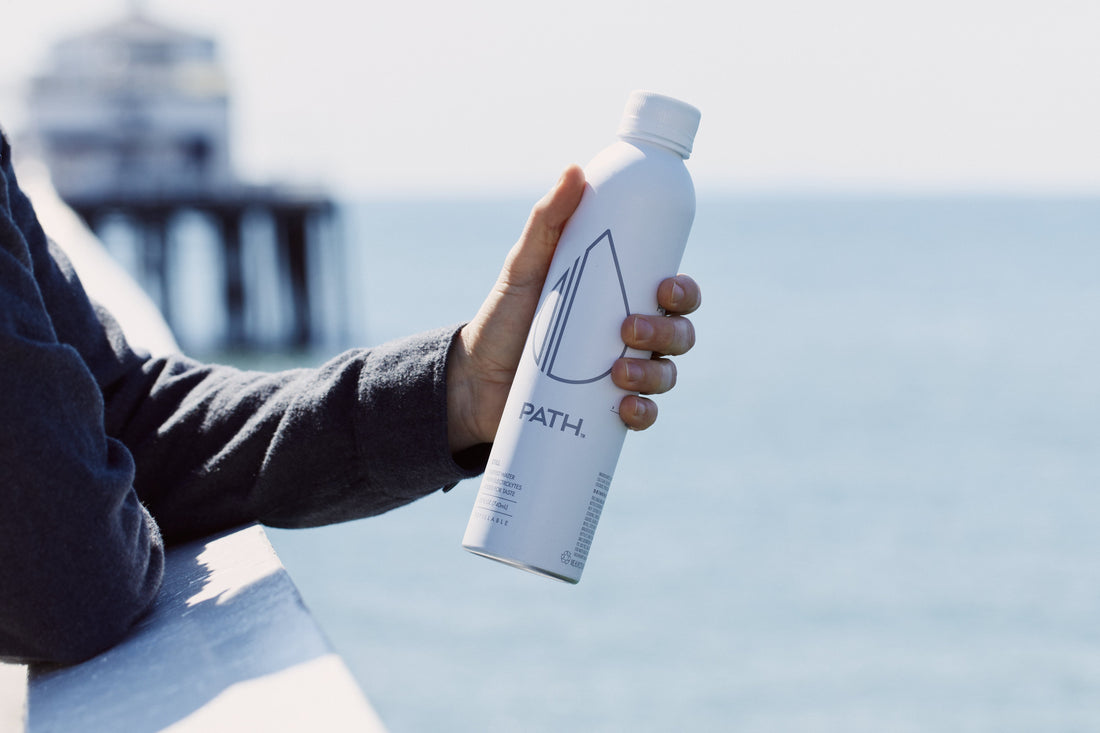 Side view of PATH aluminum bottle showcasing droplet logo and matte white finish with ocean and pier in background