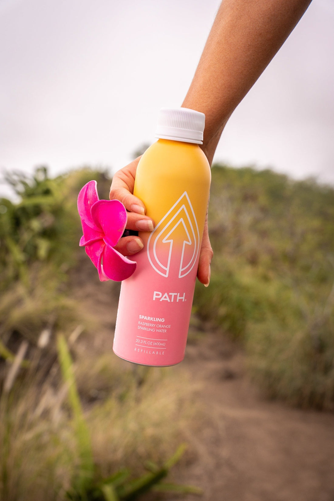 Handheld front view of PATH Raspberry Orange sparkling water bottle showing pink-to-yellow gradient, PATH logo, 'sparkling' label, and blurred beach background