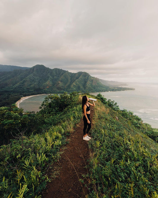 Woman drinking from a reusable water bottle while standing on a grassy coastal ridge overlooking a bay and forested mountains