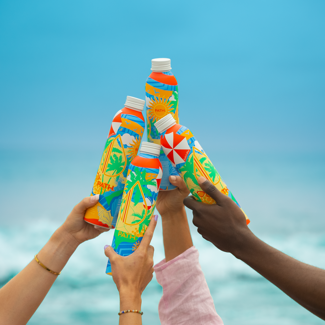 Four people clinking PATH aluminum bottled water with colorful tropical designs against a turquoise ocean backdrop