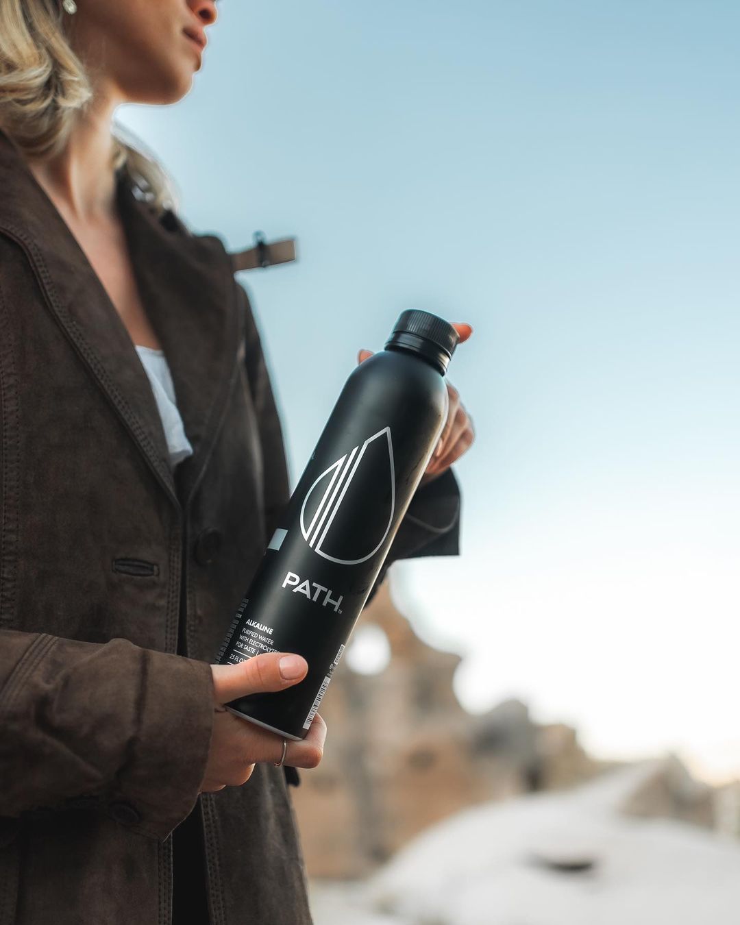 Angled view of PATH aluminum water bottle showcasing black matte finish and white logo with blurred outdoor background