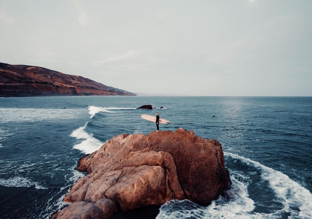 Surfer standing on a rocky outcrop holding a surfboard, looking out over the ocean under an overcast sky
