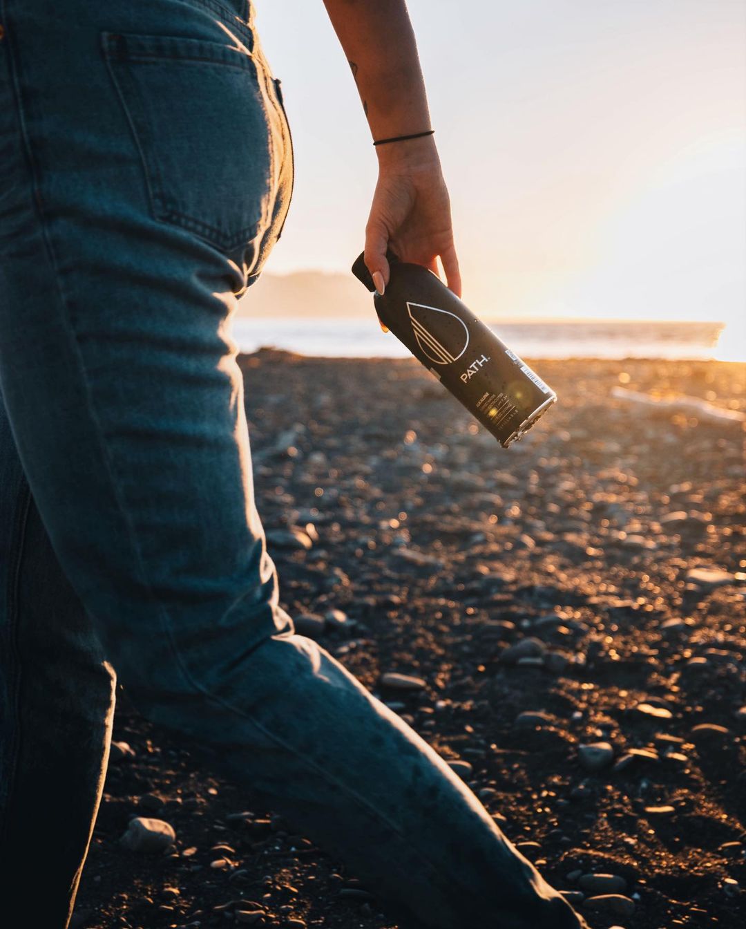 Person holding a black PATH water bottle while walking on a rocky beach at sunset