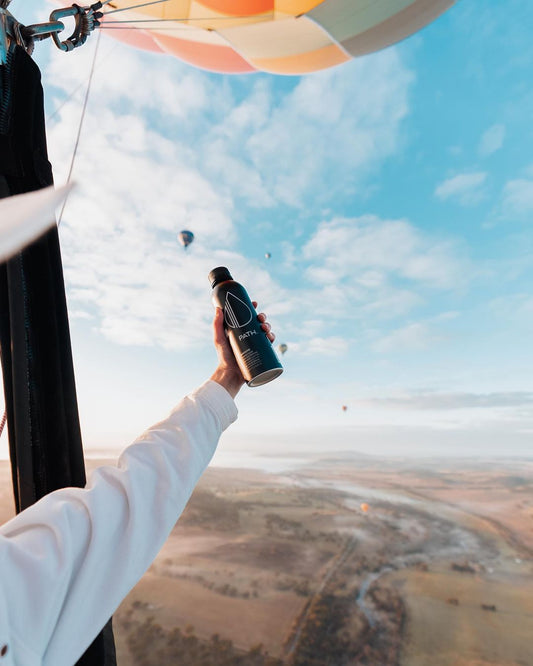 Hand holding a black PATH water bottle from a hot air balloon with rolling countryside and other hot air balloons in the sky