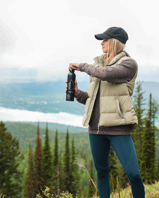 Woman opening a black PATH water bottle on a mountain overlook with evergreen trees and a lake below