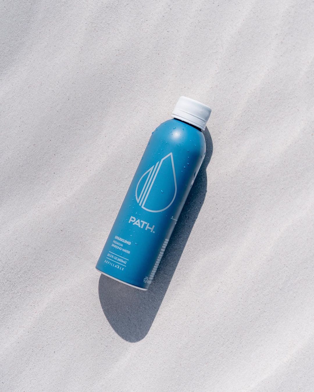 Angled view of PATH blue aluminum sparkling water bottle with white cap and water droplets on light sand
