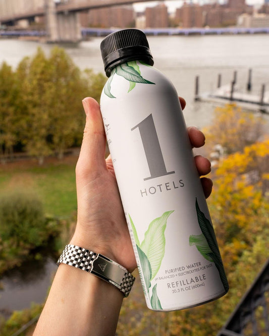 Hand holding 1 HOTELS aluminum refillable water bottle with leaf design, river and bridge in background