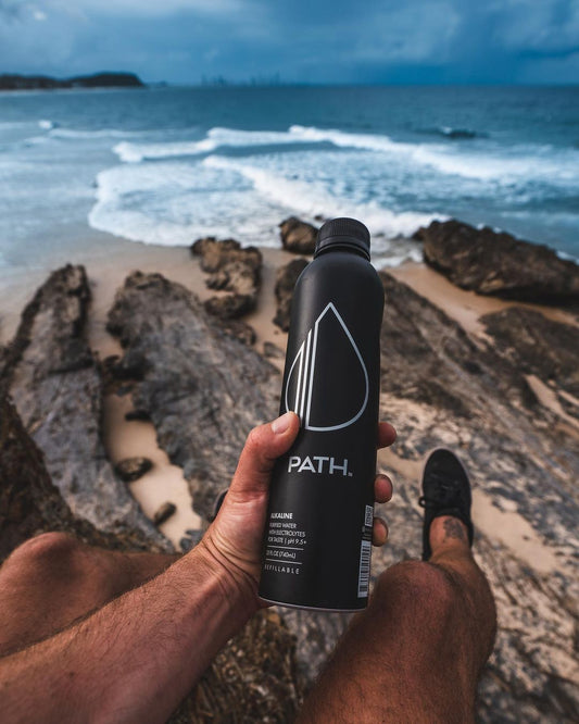 Handheld front view of PATH water bottle showing PATH logo and matte black finish with rocky shoreline and ocean waves in background