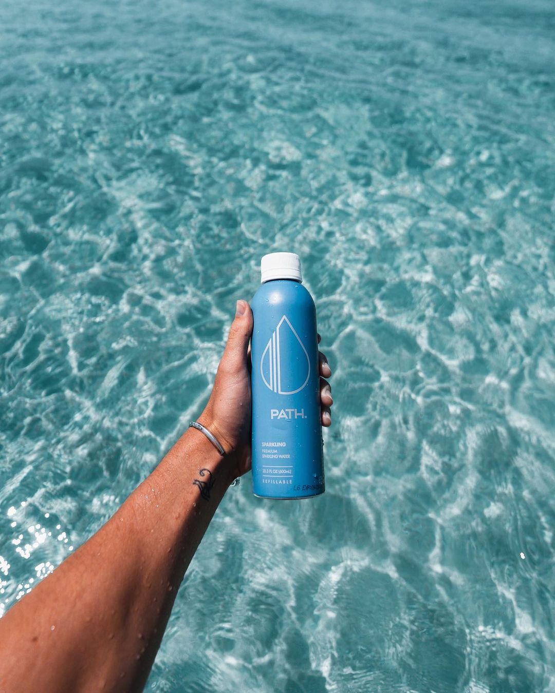 Hand holding a blue PATH water bottle over clear turquoise ocean water