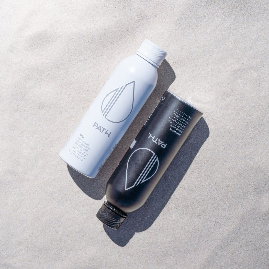 Angled top-down view of white and black PATH aluminum water bottles showing logo and caps on sand