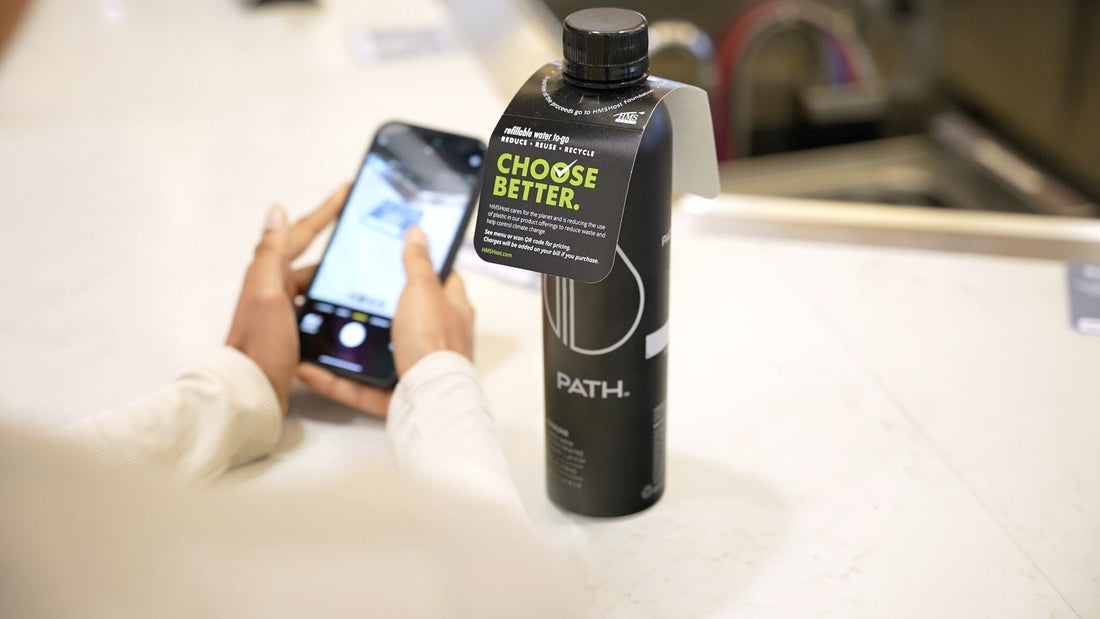 Front view of PATH aluminum bottle with CHOOSE BETTER hangtag and PATH logo on a counter, blurred hands holding a phone behind