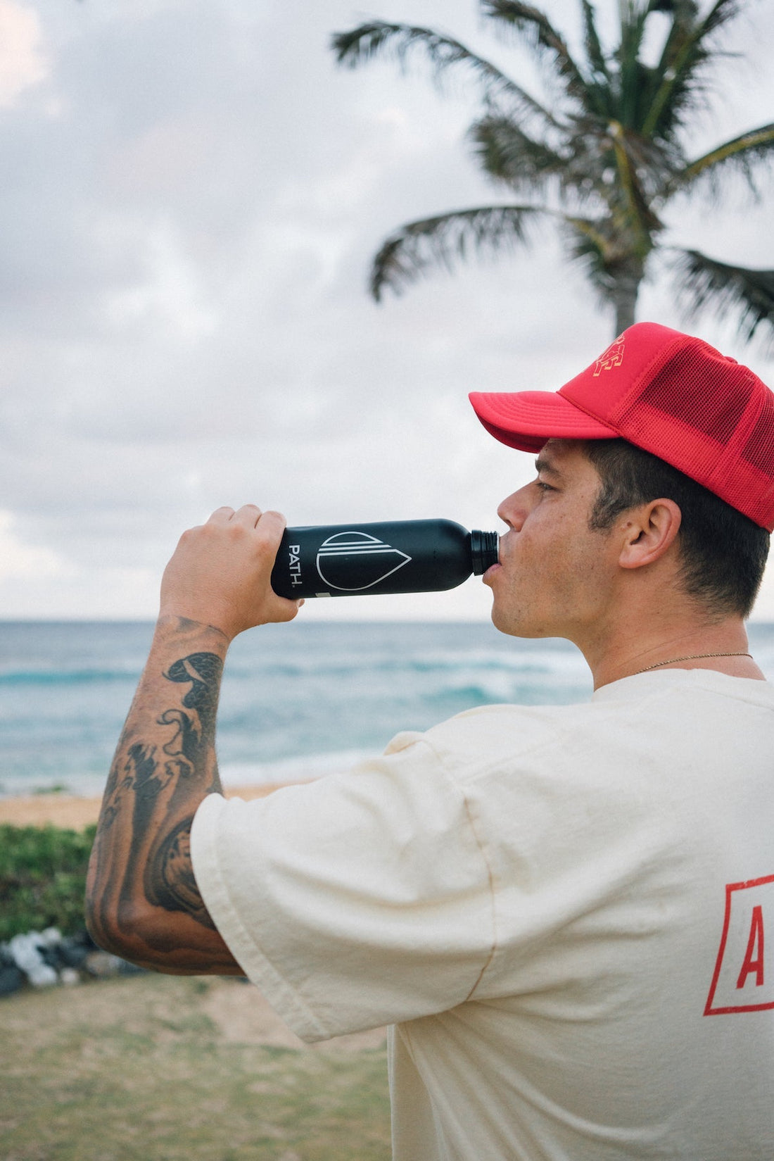 Man drinking from a black PATH reusable water bottle at a beach with a palm tree in the background