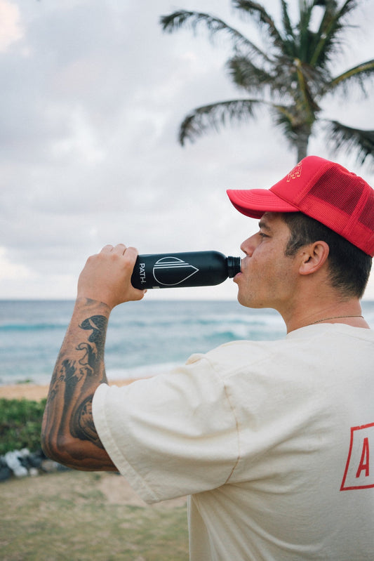Man drinking from a black PATH reusable water bottle at a beach with a palm tree in the background
